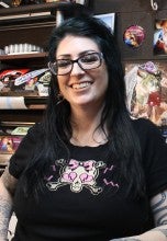 Suzen Tattoozen stands in the Whiteaker Tattoo Collective in front of a black wall with memorabilia and tools. She is wearing a black t-shirt with a skull on it.