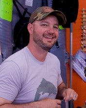 Troy Haselip sits in front of a desk with fly tying materials and an orange wall in the background. He is wearing a light gray t-shirt and a camo baseball cap.