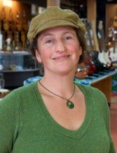 Shari Ame stands in an instrument shop with guitars in the background. She is wearing a green shirt and a light brown brimmed hat.