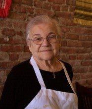 Matilda Novak stands in front of a red brick wall inside Novak's Hungarian Restaurant. She wears a black long sleeved shirt under a white apron.