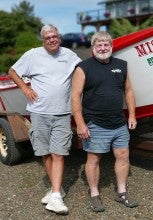Skip Bailey & David Larkins stand outdoors next to a red and white boat named "Nestucca" and a house and bushes in the background. They are both wearing shorts.