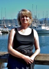 Laura Anderson stands on a pier with docked boats in the background. She wears a black tank top.