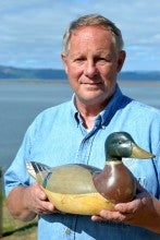 Rick Pass stands in front of a lake holding a wooden duck decoy.