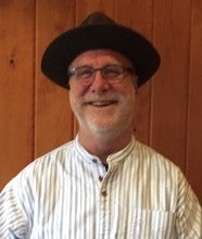 Bryan Ibach sits in front of a wood wall. He wears a black hat, a white striped shirt, and gray pants.