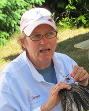 Sherry Steele sits outside with grass and bushes in the background and holds a bundle of brown feathers. She wears a white shirt and a light baseball cap.