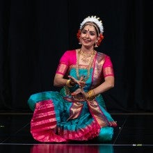 poses in an expressive dance gesture while performing on stage. She is in traditional Kuchipudi dance attire.