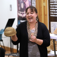 Stephanie (Wood) Craig is standing in front of an audience holding a small woven basket. She is wearing a gray shirt with black cardigan and is gesturing with her left hand.