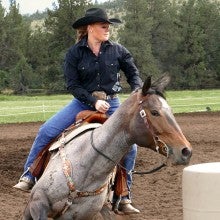 Tonya Rosebrook races around a white barrel with a gray horse. She is wearing a black long-sleeved shirt, a black cowboy hat, and blue jeans.