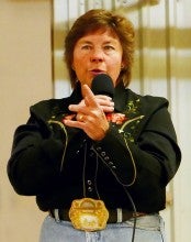 Ramona Hulick stands on a stage and talks into a microphone during an auction. She wears a black button-up shirt with gold embroidery, a large gold belt buckle, and light blue jeans.