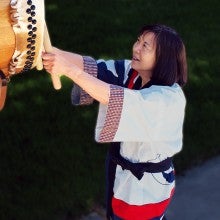 Janet Komoto stands outside with her arms outstretched and plays a taiko drum. She wears a red, white, and blue robe.