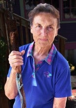 Sandy Micheli stands outside and holds a wooden stick with a green lizard on it. She wears a blue polo shirt.