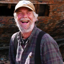the artist, Knute Nemeth, is smiling jovially at the camera. He is a middle aged man, with grey hair and a full, healthy beard. He is wearing a baseball cap, a purple and red plaid shirt, and thick black suspenders. Behind him is the brown molted hull of a boat. The sun bounces off the apples of his cheeks and the grey of his beard.