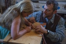 the artist, Steve Harris, kneels to the right side of the image helping his young daughter Caity with rawhide braiding. He wears a blue shirt and brown vest.