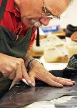Mike Strong stands over a workbench and engraves on a dark piece of leather. He wears a red shirt and gray apron.