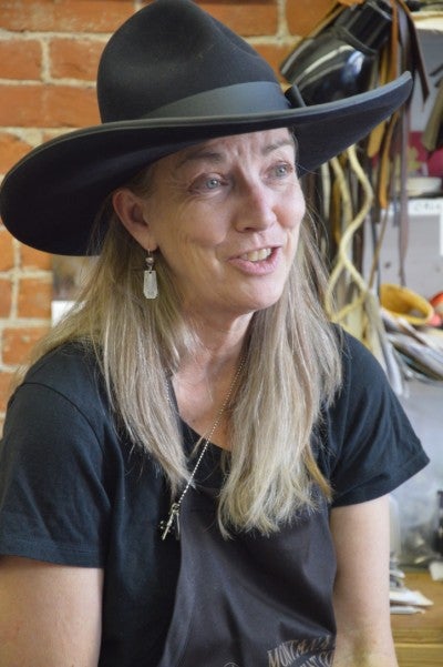 Laura sits in front of a brick wall in Montana Peaks Hat Co. and wears a black cowboy hat and black shirt.