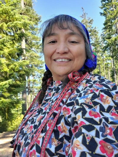 Laurie Danzuka stands in front of pine trees, wearing a shirt with a black, white, and red star design and wearing a bandana over her braided hair.