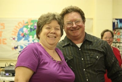 Sarge and CeCe Glidewell stand together inside a room with cream walls. CeCe is wearing a purple shirt and Sarge is wearing a black striped button-up shirt. 