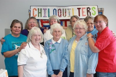 Group photo of the Chiloquilters (eight people) standing in front of a white banner that reads "CHILOQUILTERS" in rainbow letters.