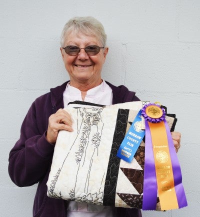 Cheryl Hobbs holds an award-winning quilt in front of a white wall of the Northwestern Motel in Heppner, Oregon. She wears a purple sweatshirt over a white collared shirt.