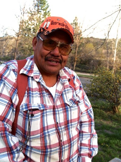Larry Dick stands outside and wears a plaid shirt, orange hat, and sunglasses.