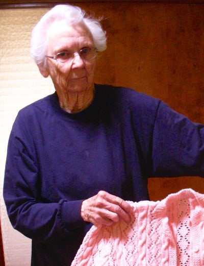 Emma Jean Smith stands indoors holding up a light pink knitted cardigan. She wears a dark blue sweater.