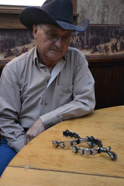 Larry Bacon sits at a table with a pair of silver spurs and wears a blue shirt and blue cowboy hat.