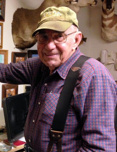 Donald Webb stands in his home in Vernonia, Oregon with framed artwork and taxidermy on the walls behind him. He wears a purple collared shirt with black suspenders and a camo baseball cap.
