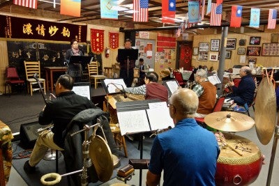 Yat Sing Music Club practice together, holding a variety of instruments including strings, prass, and percussion. Two people stand at the front of the room in front of music stands.