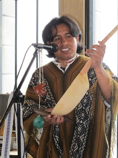  Alex Llumiquinga Perez stands in front of a microphone and displays a charango. He is wearing a brown, black, and white patterned poncho. 