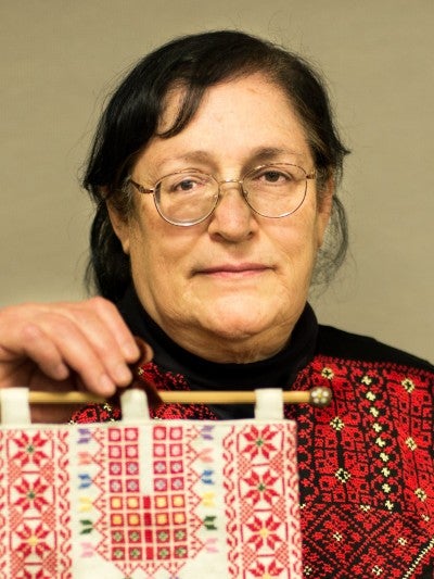 Feryal Abbasi-Ghnaim stands against a white wall holding up a piece of red and white embroidery. She wears a red and black embroidered outfit.