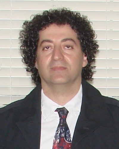 Houshang Sedighi stands against a white background. He wears a black suit and a red and black tie.