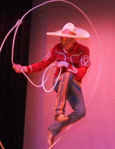 Jose Antonio Huerta demonstrates rope work on stage. He wears a red collared shirt, blue jeans, cowboy boots and a white cowboy hat.