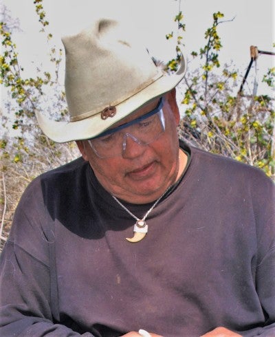 Cecil Coons sits on a stump outside in Burns, Oregon and demonstrates how to chip an arrowhead (obsidian). He wears a black long sleeved shirt, a white cowboy hat, and a talon pendant. 