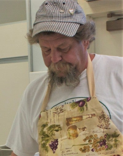 Wally Cunial stands in a kitchen and prepares traditional Italian food. He wears a white t-shirt, a cream printed apron, and a checkered baseball cap.