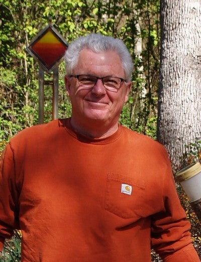 Tom Herrera stands outdoors next to a welded sculpture. He is wearing a rust colored shirt and blue jeans.