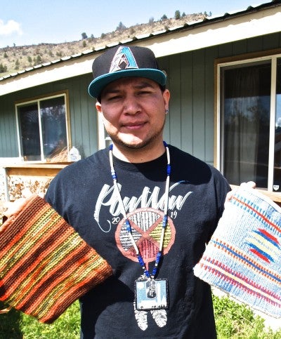 Jefferson Greene stands in front of a house and holds two colorful baskets. He wears a black t-shirt that says "Powwow 2019" and a black hat with a decorated letter "A".
