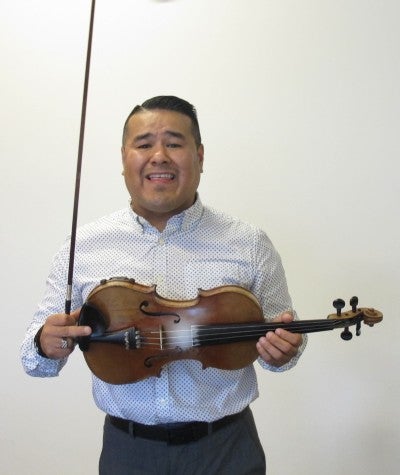 José Ramirez stands in front of a white wall holding a violin and wearing a white collared shirt.