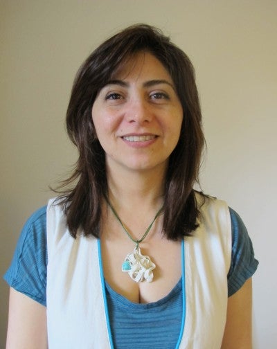 Marjan Anvari stands in front of a white wall and wears a blue t-shirt, a white vest, and a blue and white pendant.
