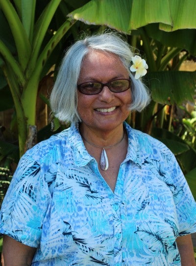 Lei McCornack stands outside in front of banana palms. She wears a blue collared shirt with palm fronds printed on it, and a yellow and white tropical flower tucked behind her left ear.