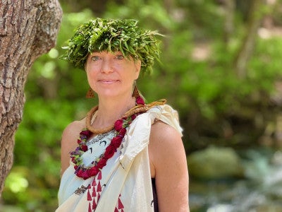 Andrea Luchese is wearing a cream dress with red and black patterns, as well as a rose lei. She is standing in front of a forest.
