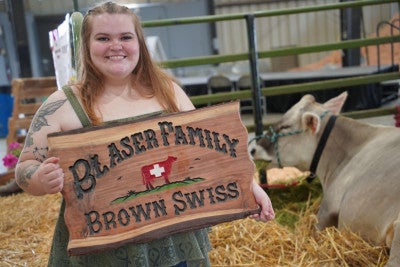 Clara Blaser holding a wooden sign saying Blaser Family Brown Swiss in a barn with a cow behind her. 