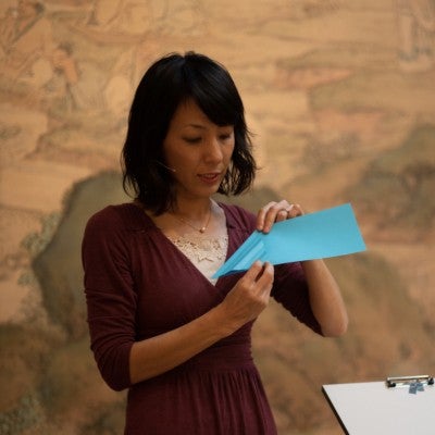 A Japanese woman wearing a plum colored blouse folds a blue piece of paper