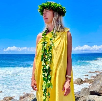 Andrea Luchese wearing a yellow dress and leaf leis. She is standing outside on the rocks with the ocean behind her.