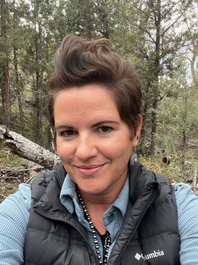 Clair Kehrberg in a blue shirt and black vest, with a forest in the background