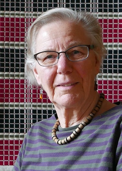 Leena Mela Riker sits in front of a woven hanging blanket with a a white, black, and red striped plaid design. She wears a grey and black striped long sleeve shirt.