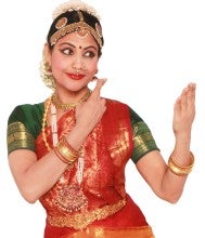Jayanthi Raman stands in an expressive dance pose against a white background. She wears a red, green, and gold silk sari and beaded ornamental headwear.