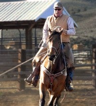 Dan Fulwyler practices roping on a brown mustang outside at his home arena. He wears a white long sleeved shirt under a tan vest, dark brown leather chaps, and a camo baseball hat. 