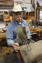 Dave Clowes stands in front of a tree (supportive structure for a saddle) and holds a knife and a brand in his western shop in Bonanza, Oregon. He wears a brown apron over a blue collared shirt, and a white cowboy hat. 