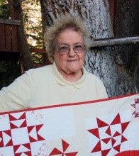 Betty Morris stands outside in front of a tree and holds a red and white winter themed quilt. She wears a pale yellow shirt.