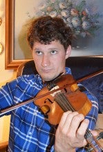 Eric Jepsen sits in a chair in front of a framed flower painting playing a fiddle. He wears a blue plaid shirt.